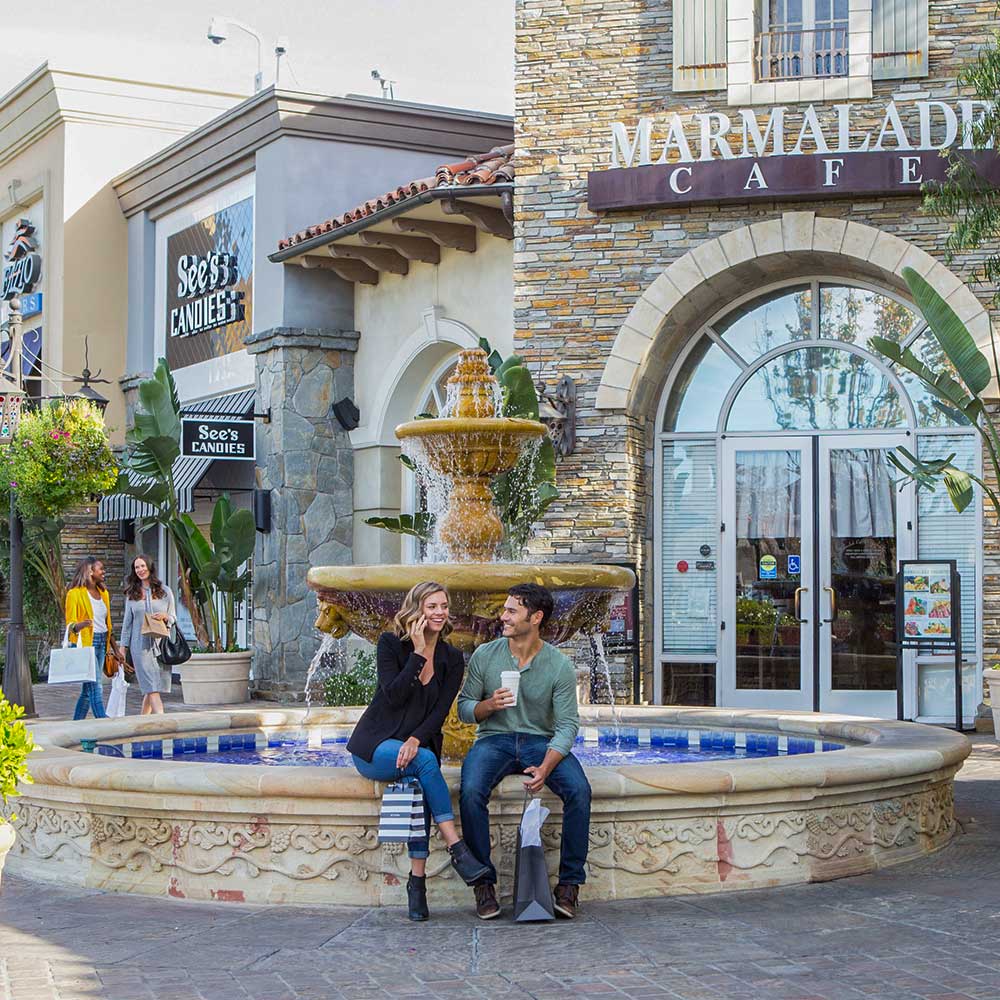 people sitting by a decorative fountain outside marmalade cafe and see's candies at an outdoor shopping center